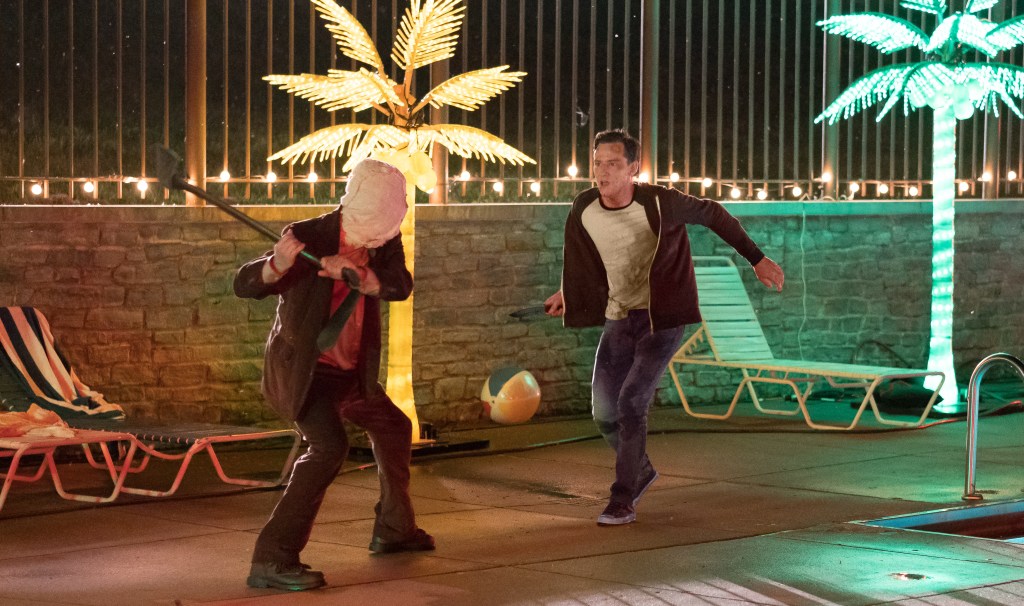 An image from the film The Strangers: Prey at Night. A masked man is swimming an axe at another man (Bill Pullman) by the edge of a swimming pool. There are neon palm trees and sun loungers in the background.
