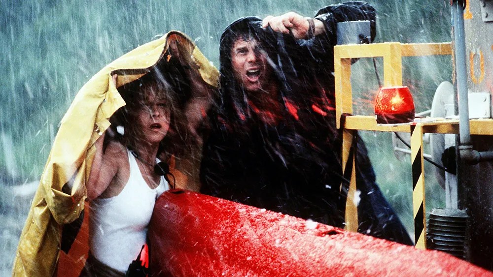 An image from the film Twister. It features a man (Bill Paxton) and a woman (Helen Hunt) in a hailstorm. They are standing at the back of a red truck and are both using rain jackets to shelter from the storm. 