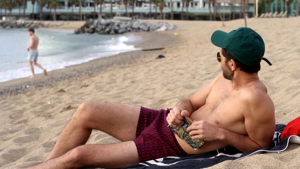 An image from the film End of the Century. It features a man (Juan Barberini) lying on a beach holding a book. He's looking at another man (Ramon Pujol) who is walking into the water further along the beach. 