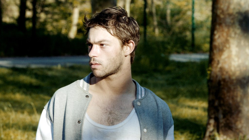 An image from the film Sauvage. It features a man (Félix Maritaud). He is standing on the side of a road looking off to the side. 