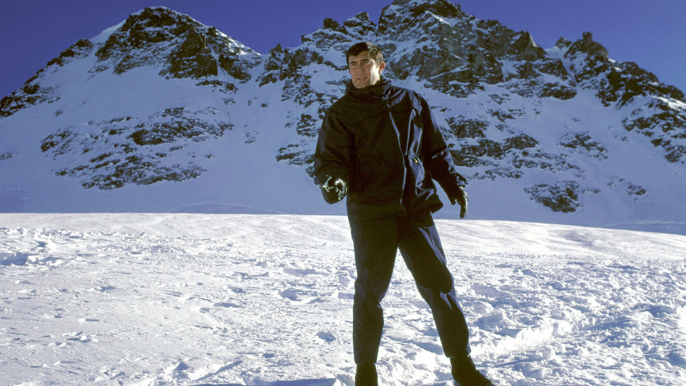 An image from the film On Her Majesty's Secret Service. It features a man (George Lazenby) standing on a snowy mountain. He's holding a pistol and pointing it in front of him.