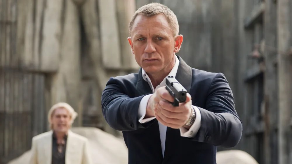 An image from the film Skyfall, it features a man (Daniel Craig) holding a pistol. He is holding it with both hands and pointing it downwards at something in front of him. Another man (Javier Bardem) can be seen in the background. 