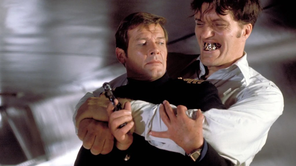 An image from the film The Spy Who Loved Me. It features two men (Roger Moore and Richard Kiel). One man has his arms wrapped around the other and has large silver teeth. 