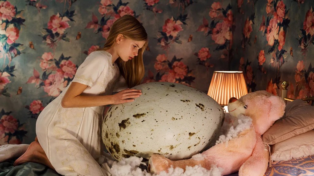 An image from the film Hatching. It features a young girl (Siiri Solalinna) on top of a bed with a very large egg. 