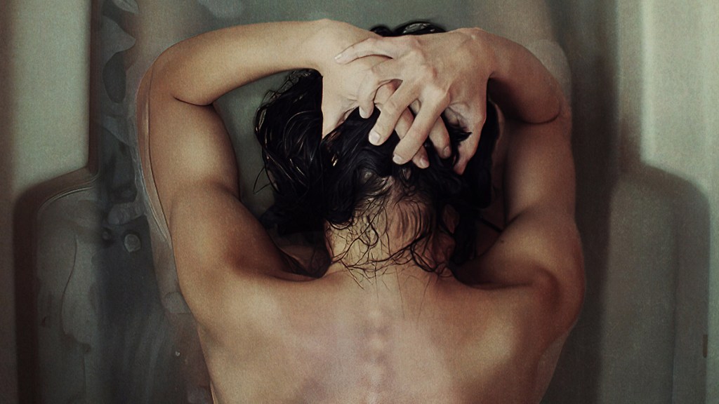 An image from the film Huesera: The Bone Woman. It features an overhead shot of a woman in the bath. She has her hands on her head and is crouched over. The outline of her spine can be seen through her back. 