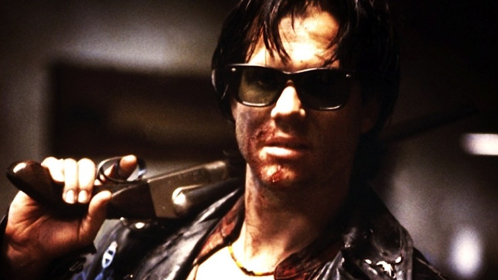 An image from the film Near Dark. It features a man (Bill Paxton). He has cuts on his face and is wearing dark sunglasses. He's also holding a gun and has it resting over his shoulder. 