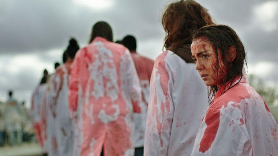 An image from the film Raw. It features a young woman (Garance Marillier) at the back of a line of other young people. They are white wearing overalls stained with red paint. 