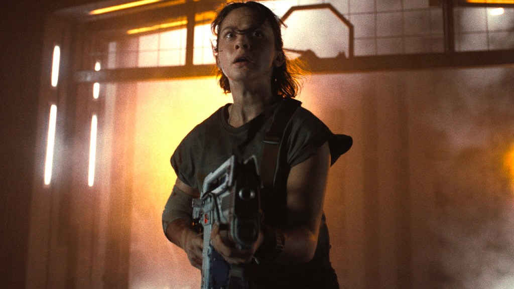 An image from the film Alien Romulus. It features a woman (Caliee Spaeny) standing in front of a large elevator shaft holding a gun.