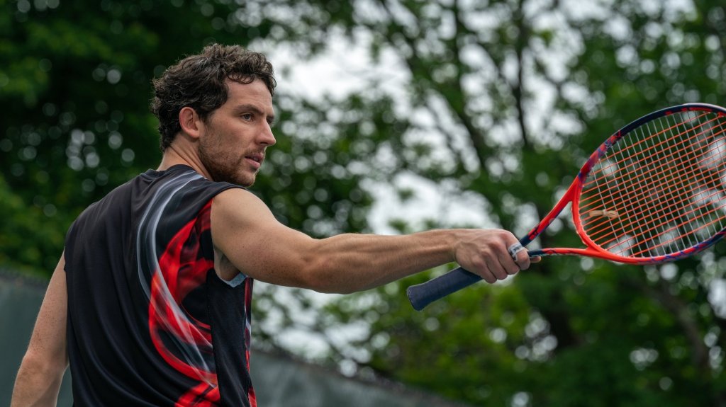 An image from the film Challengers. It features a man (Josh O’Connor) playing tennis. He is holding a racquet and is pointing it in front of him.