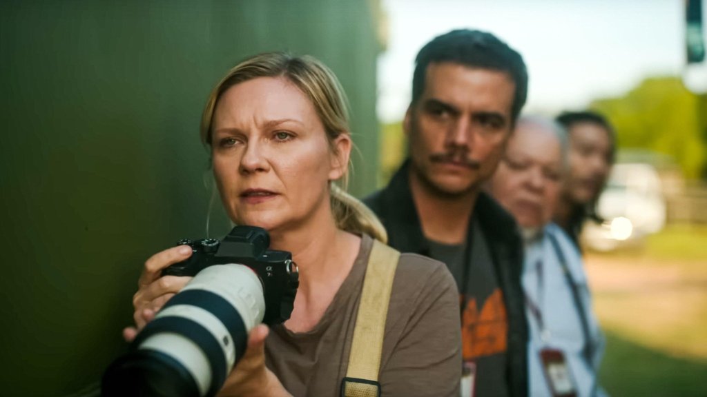 An image from the film Civil War. It features a woman (Kirsten Dunst) standing by the side of a building with a camera in her hands. Three men are standing behind her.