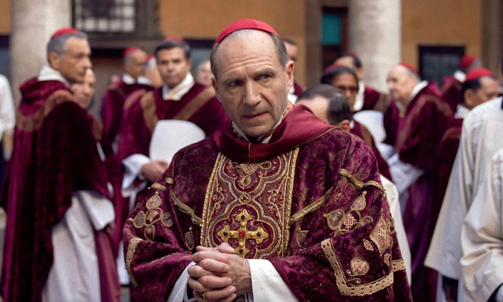 An image from the film Conclave. It features a Cardinal (Ralph Fiennes) standing in front of a large crowd of other Cardinals.