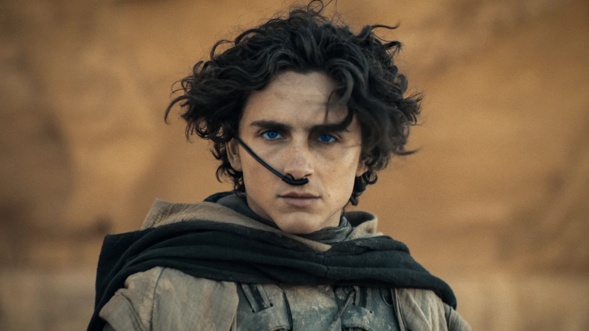 An image from the film Dune Part II. It features a man (Timothée Chalamet) in a desert setting. He has a black tube running from his nose to behind his ear.