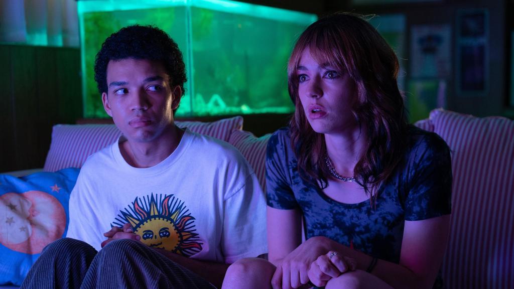 An image from the film I Saw the TV Glow. It features a man (Justice Smith) and a non-binary person (Brigette Lundy-Paine) sitting on a sofa, a purple glow from the TV can be seen in the room.