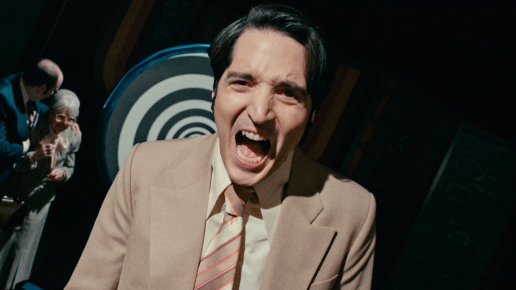 An image from the film Late Night With the Devil. It features a man (David Dastmalchian) screaming with a black and white spiral behind him.