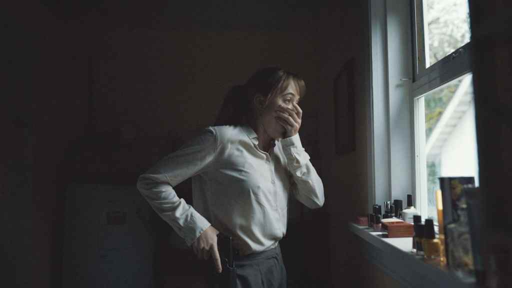 An image from the film Longlegs. It features a woman (Maika Monroe) looking out of a window and holding her hand over her mouth in shock.