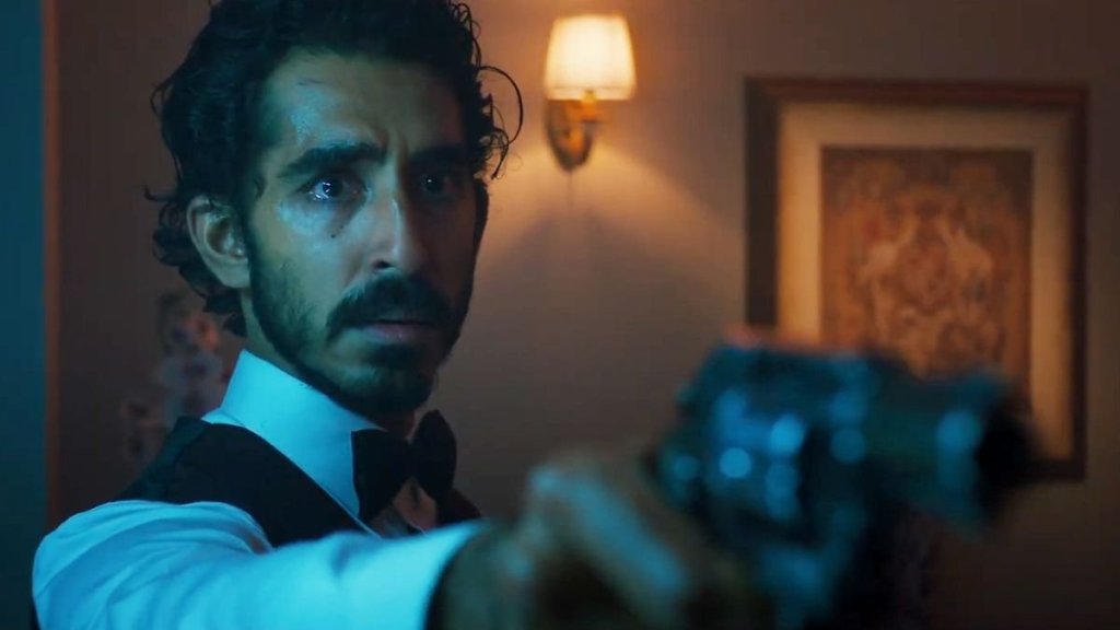 An image from the film Monkey Man. It features a man (Dev Patel) holding a gun out in front of him. He’s wearing a waiter’s uniform.