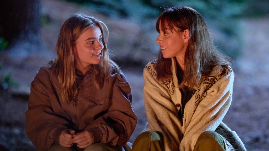 An image from the film My Old Ass. It features two women (Maisy Stella and Aubrey Plaza) sitting on a log looking at each other.