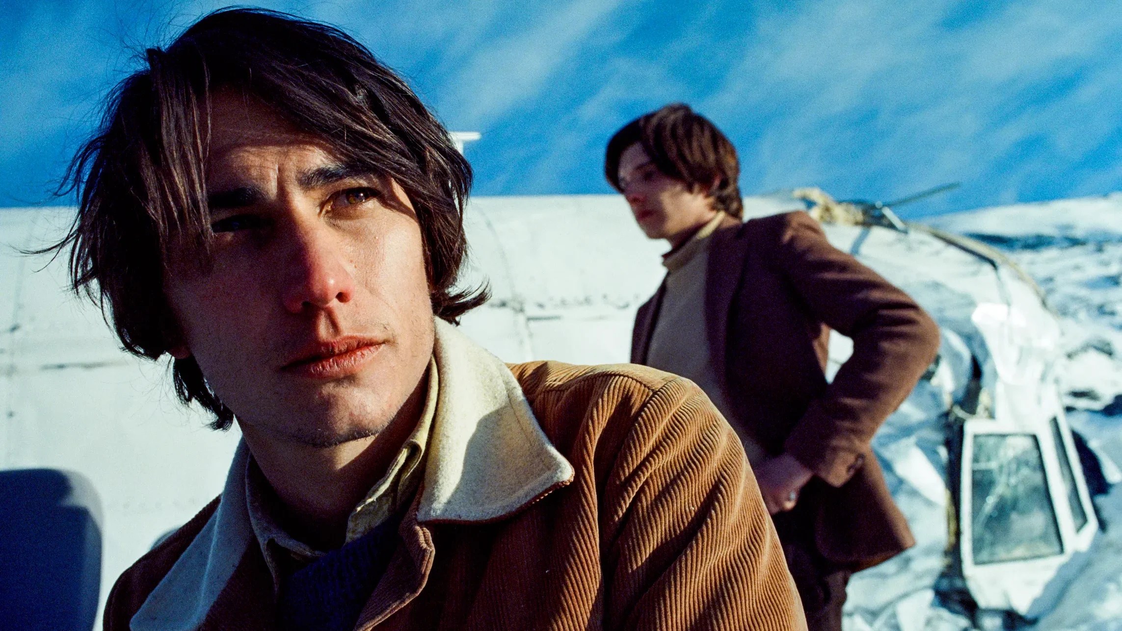 An image from the film Society of the Snow. It features two men standing in front of a plane wreckage on a snowy mountain.