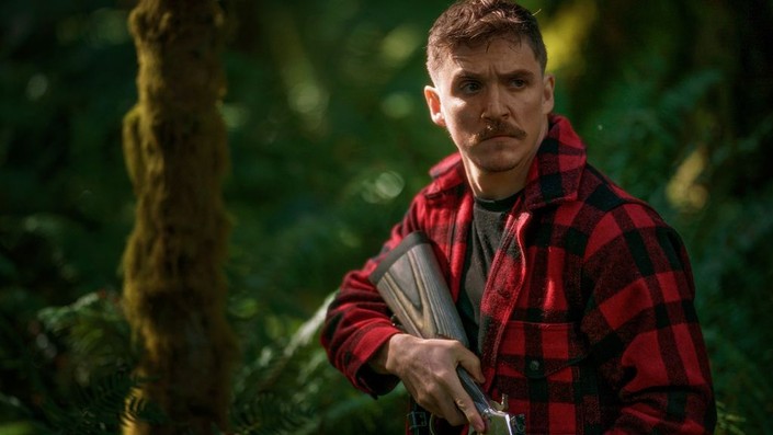 An image from the film Strange Darling. It features a man (Kyle Gallner) in a forest holding a shotgun.