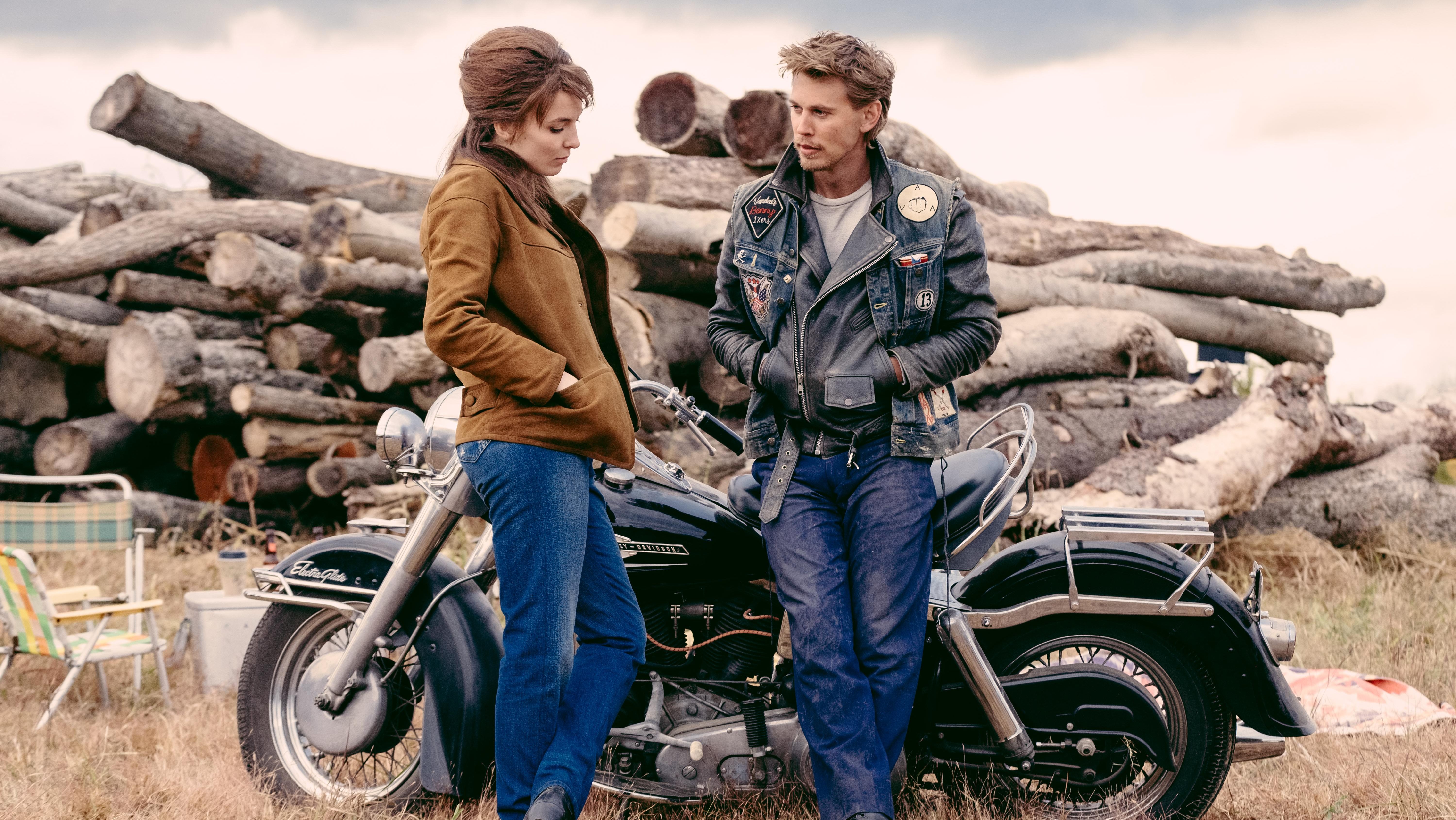 An image from the film The Bikeriders. It features a man (Austin Butler) and a woman (Jodie Comer) standing in front of a motorcycle.