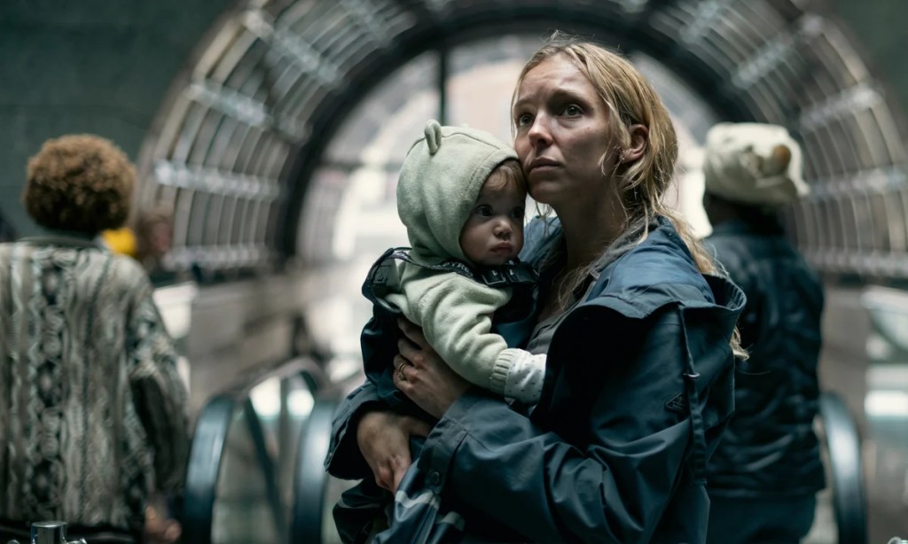 An image from the film The End We Start From. It features a woman (Jodie Comer) holding a baby at the top of a set of escalators.