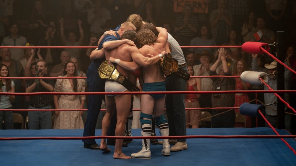 An image from the film The Iron Claw. It features a group of five men in a huddle in the middle of a wrestling ring.