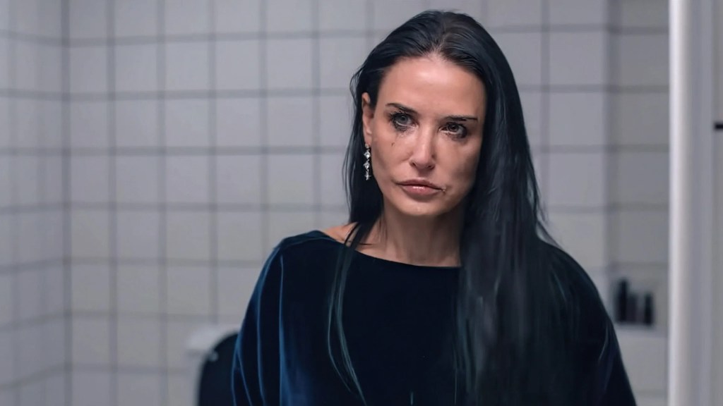 An image from the film The Substance. It features a woman (Demi Moore) looking at herself in a bathroom mirror. Her makeup is slightly smudged and she looks sad.