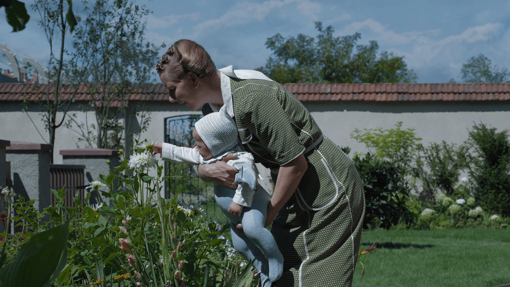 An image from the film The Zone of Interest. It features a woman (Sandra Huller) holding a baby in front of some plants in a large garden.