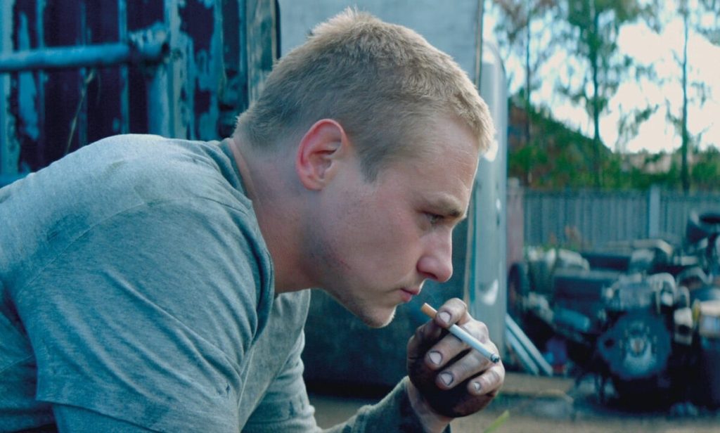 An image from the film Unicorns. It features a man (Ben Hardy) sitting outside a garage smoking.