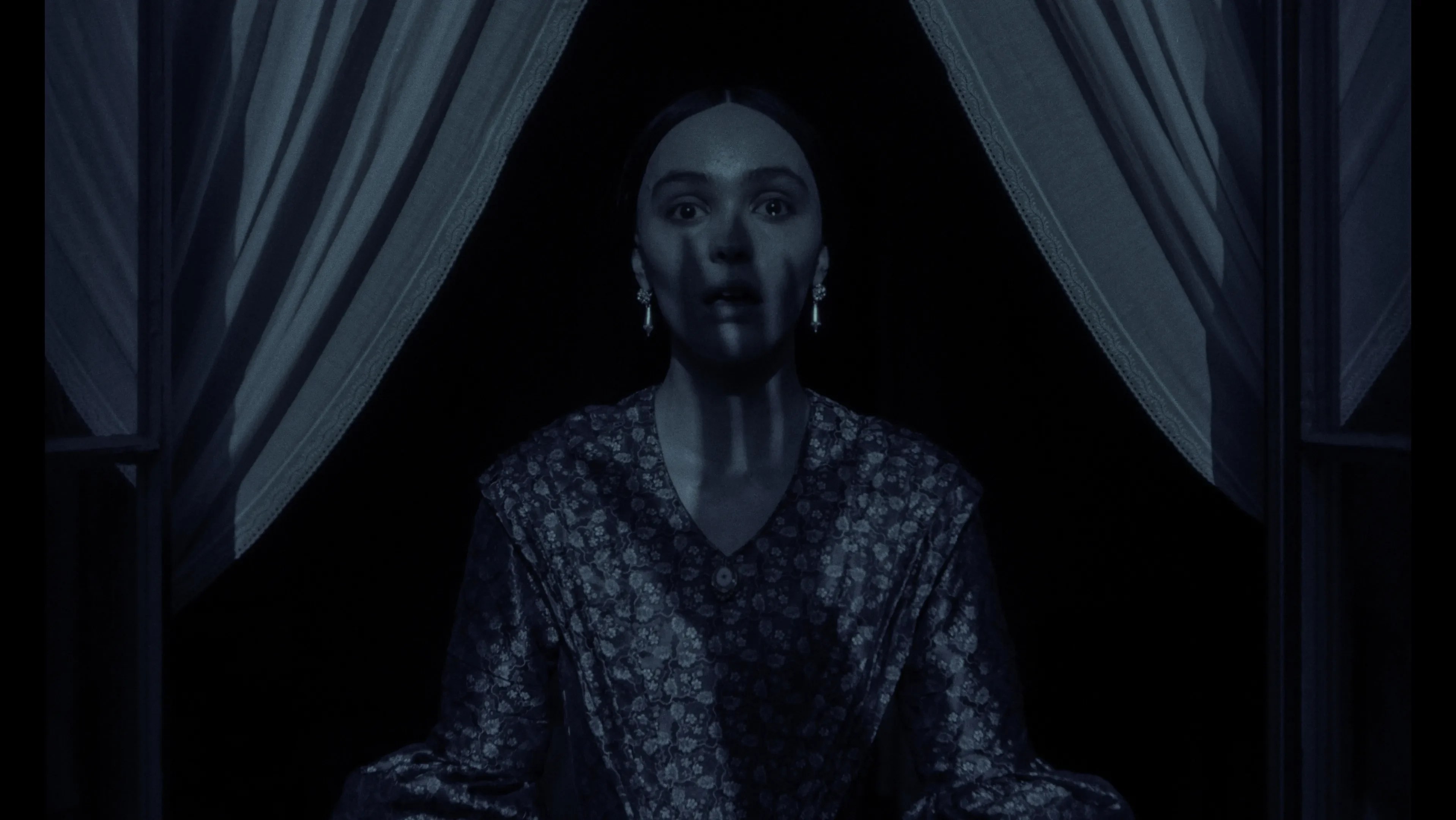 An image from the film Nosferatu. It features a woman (Lily-Rose Depp) standing at a window. The shadow of a large hand can be seen over her face. 