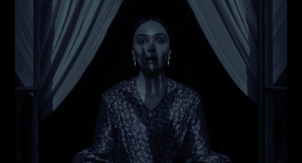 An image from the film Nosferatu. It features a woman (Lily-Rose Depp) standing at a window. The shadow of a large hand can be seen over her face. 