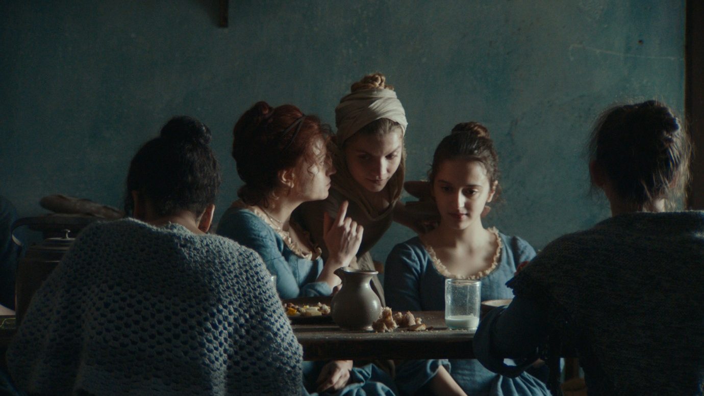 An image from the film Gloria! It features a group of young women sitting at a table. One of the women is whispering something to a young maid who is crouching down to listen. 