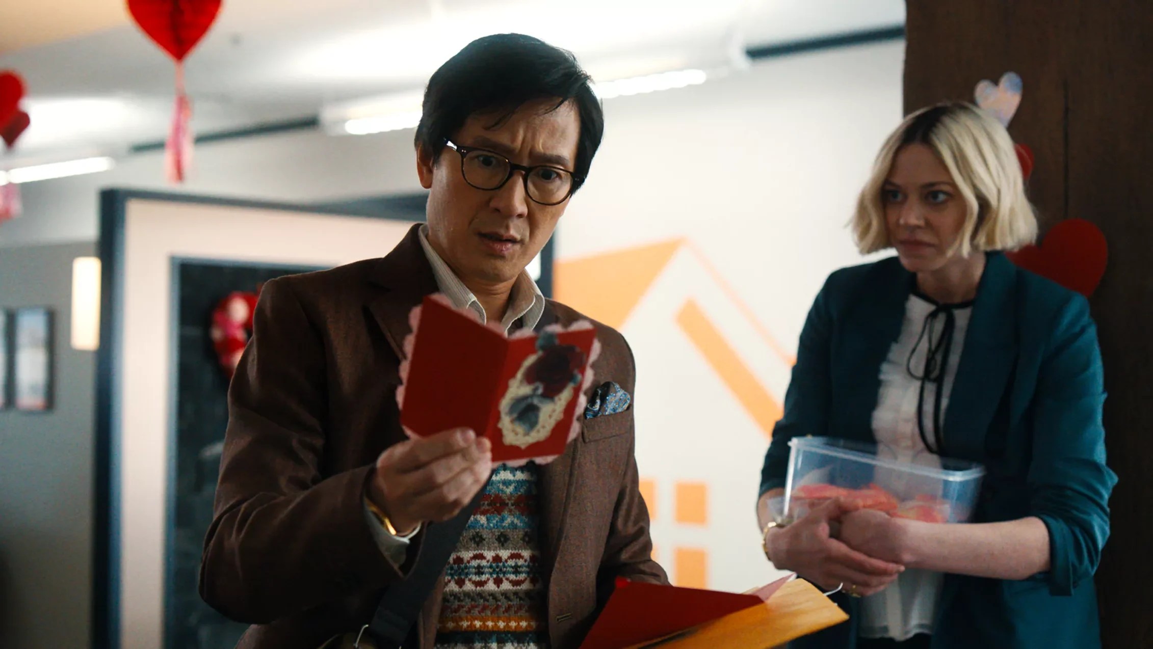 An image from the film Love Hurts. It features a man (Ke Huy Quan) reading a Valentine’s Day card in an office. A woman (Lio Tipton) is standing beside him and watching his reaction. 