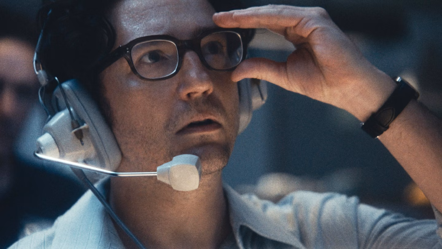 An image from the film September 5. It features a man (John Magaro) wearing a large set of headphones with a microphone attached. He has one hand on his glasses. 