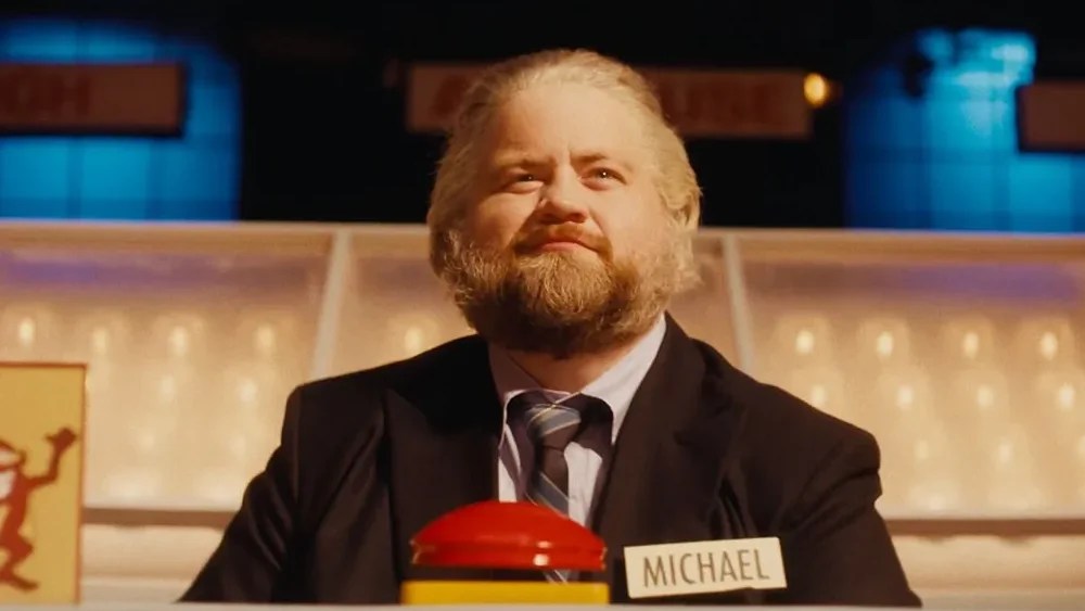 An image from the film The Luckiest Man in America. It features a man (Paul Walter Hauser) sitting behind a large red buzzer on the set of a TV gameshow.