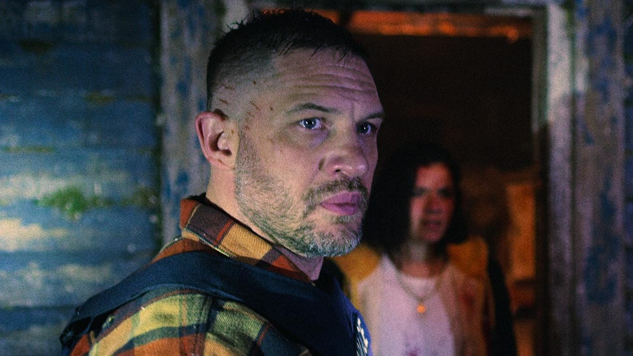 An image from the film Havoc. It features a man (Tom Hardy) standing in front of a cabin. He's wearing a police vest and has blood stains on his face. 
