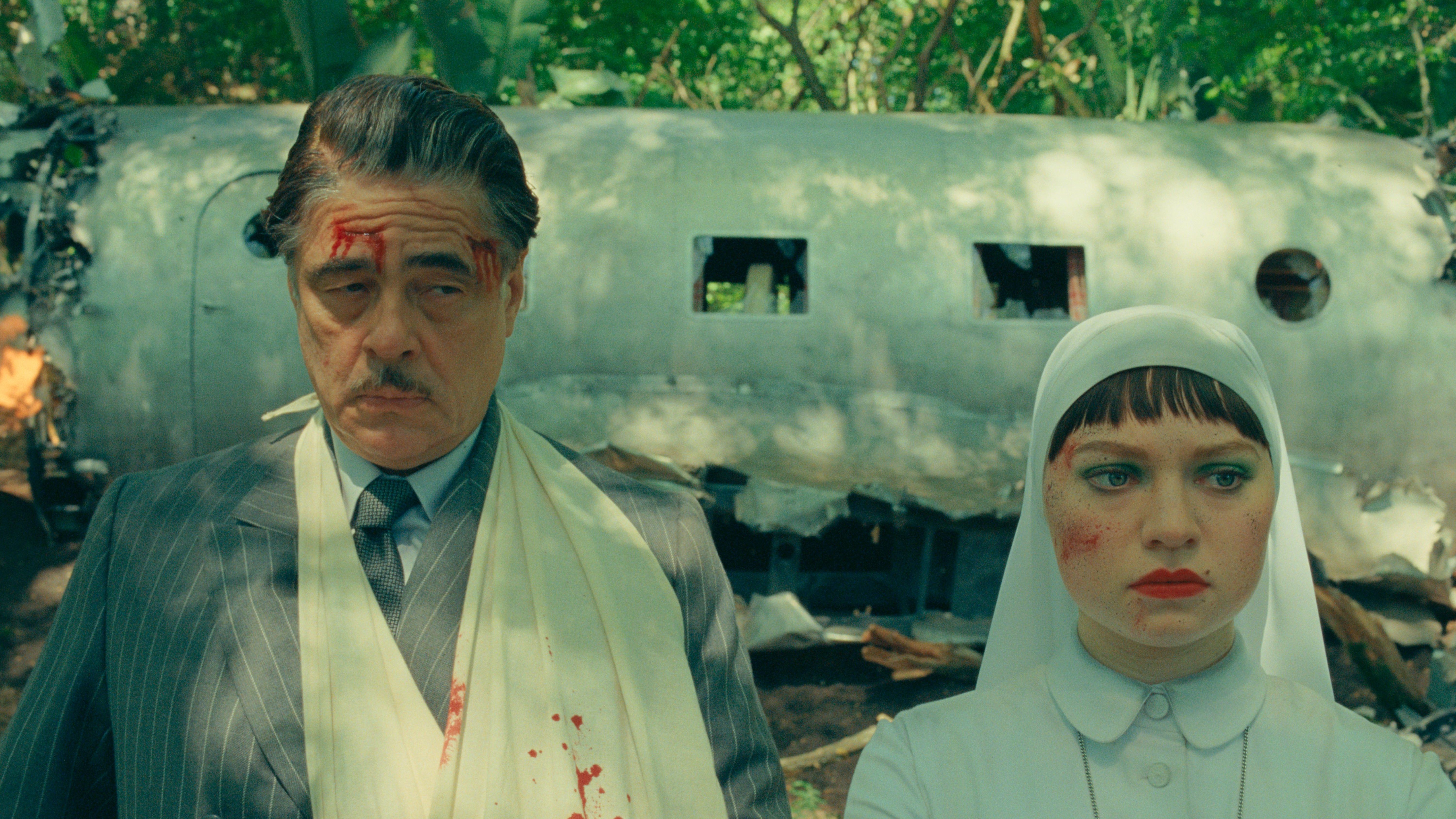 An image from the film The Phoenician Scheme. It features a man (Benicio del Toro) and a woman (Mia Threapleton) standing in front of a plane wreckage.