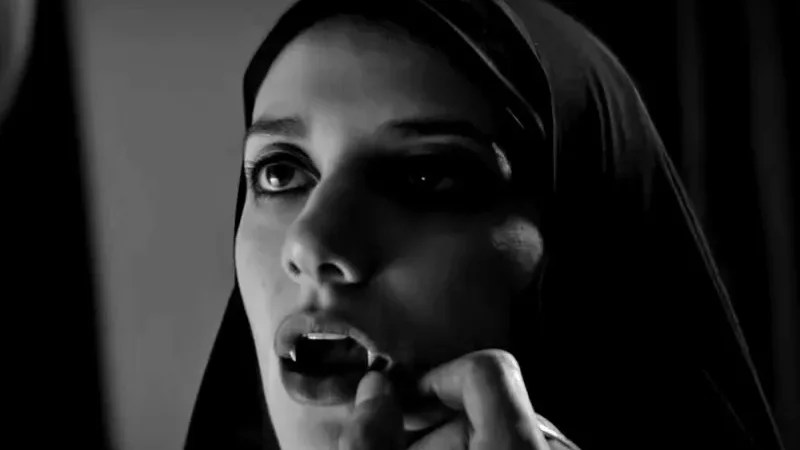 An image from the film A Girl Walks Home at Night. It features a female vampire (Sheila Vand). She has her mouth open exposing her fangs while the hand of someone in front of her feels them. 