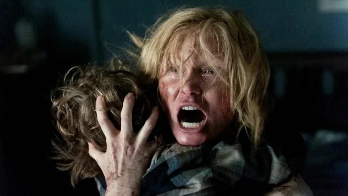 An image from the film The Babadook. It features a woman (Essie Davis) screaming. She is holding her young son (Noah Wiseman) against her with his head faced the opposite direction to hers and holding it with her hand. 
