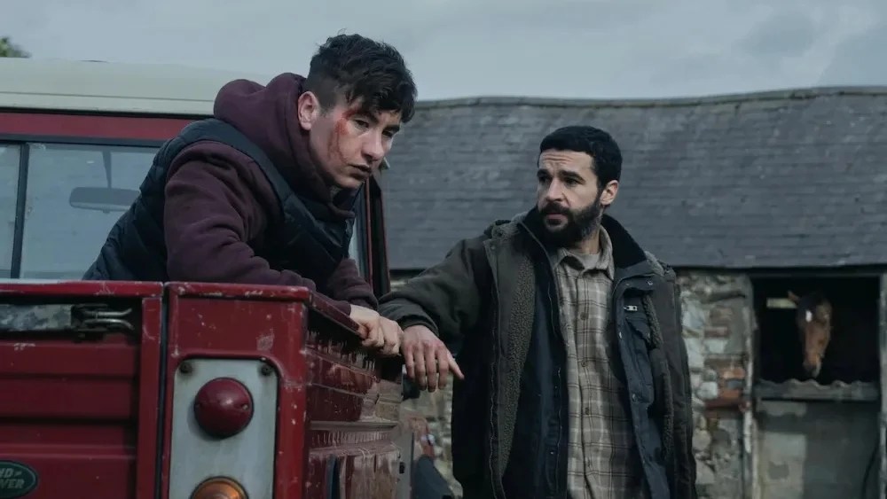 An image from the film Bring Them Down. It features two men. One (Barry Keoghan) is sitting in the back of a truck and has blood stains on his face while the other (Christopher Abbott) is leaning against the side of the same truck.