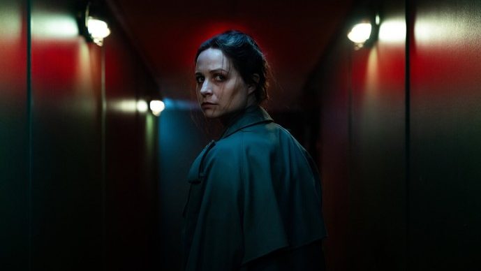 An image from the film Censor. It features a woman (Niamh Algar) standing in a corridor. She has her head turned looking behind her. 