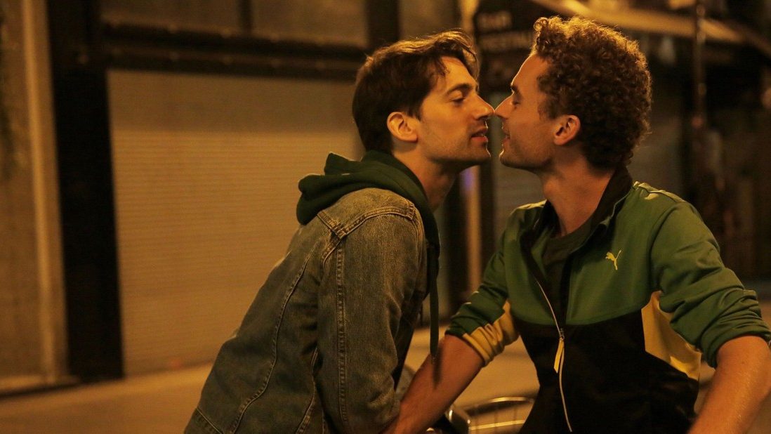 An image from the film Paris 05:59: Théo & Hugo. It features two men (Geoffrey Couët and François Nambot) riding bikes through city streets at night. They are stationary and leaning in to each other to kiss. 