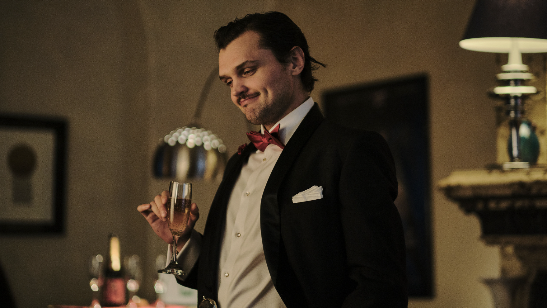 An image from the film Borderline. It features a man (Ray Nicholson) dressed in a black tuxedo with a red bowtie. He's holding a glass of champagne. 