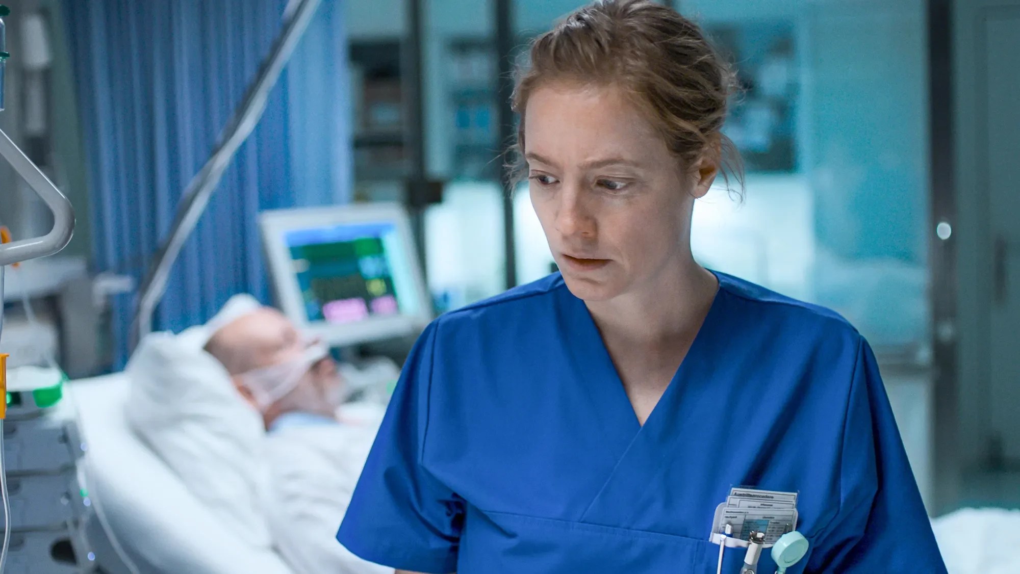 An image from the film Late Shift. It features a nurse (Leonie Benesch) in a hospital. A male patient can be seen in a bed behind her.