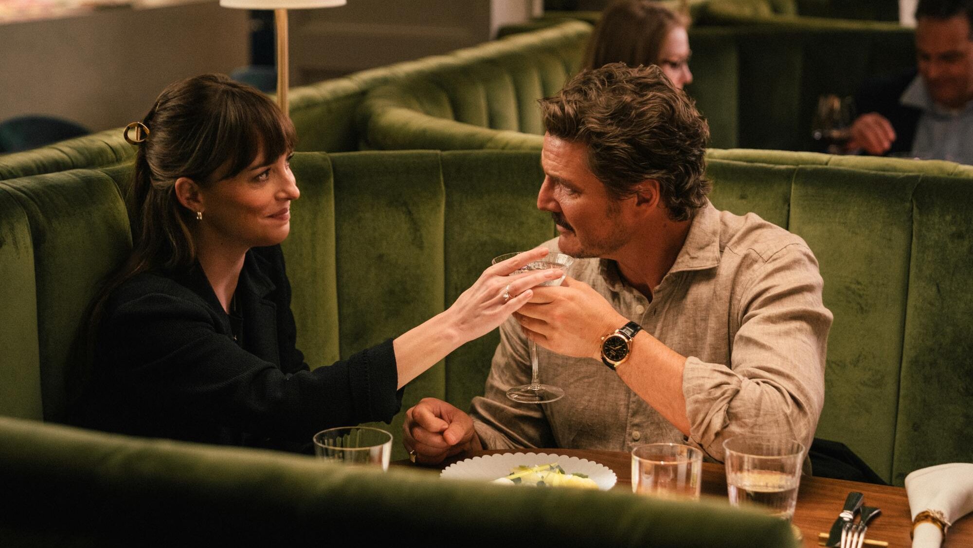 An image from the film Materialists. It features a man (Pedro Pascal) and a woman (Dakota Johnson) sitting at a table in a restaurant. The man is holding a drink and the woman is taking it off him while they look at each other.