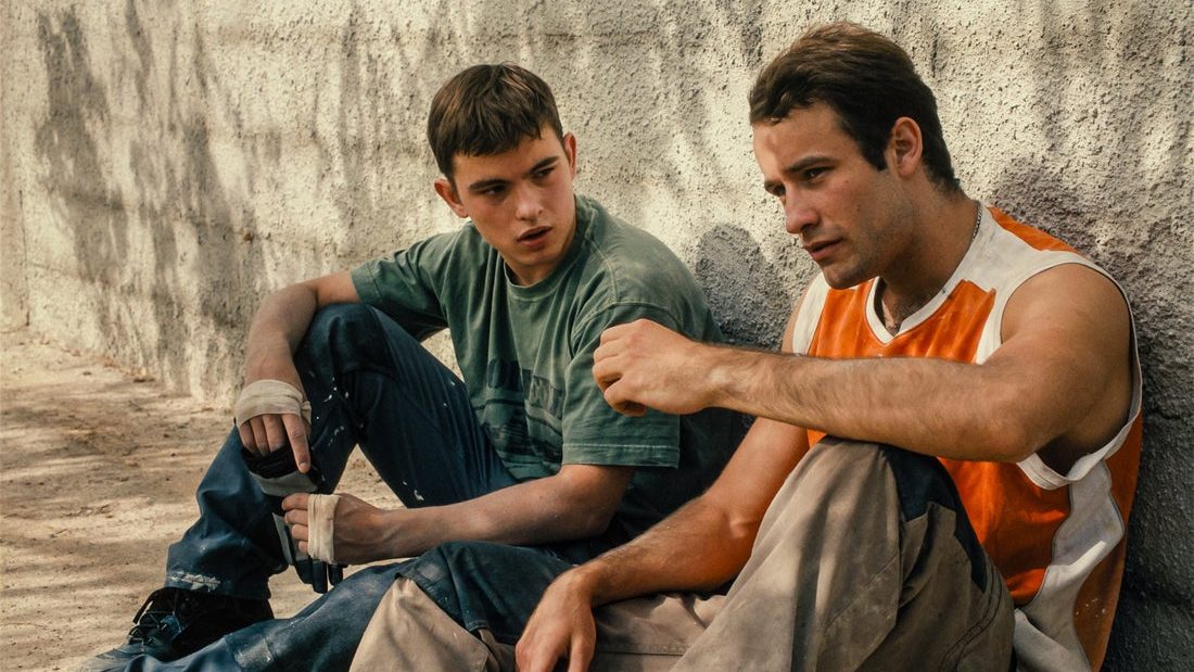An image from the film Enzo. It features an image of a teenage boy (Eloy Pohu) and a man (Maksym Slivinskyi) sitting against a wall. 