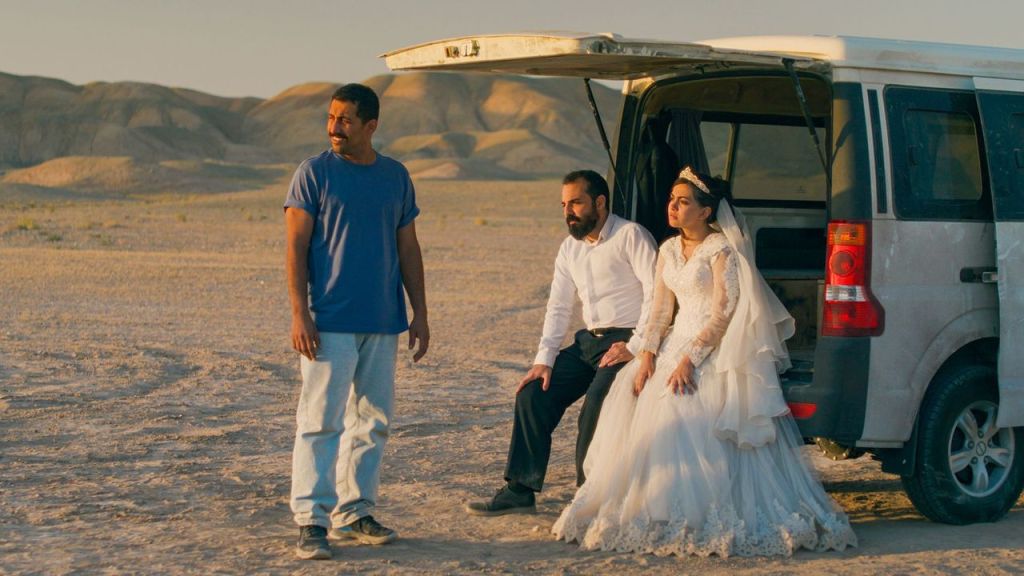 An image from the film It Was Just an Accident. It features two men and a woman wearing a wedding dress in a desert sitting on the boot of a large car. 