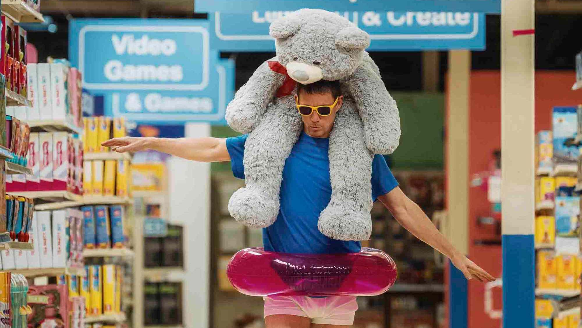 An image from the film Roofman. It features a man (Channing Tatum) skating through a toy store on heelys. He has a pink rubber ring around his waist and a large teddy bear on his shoulders. 