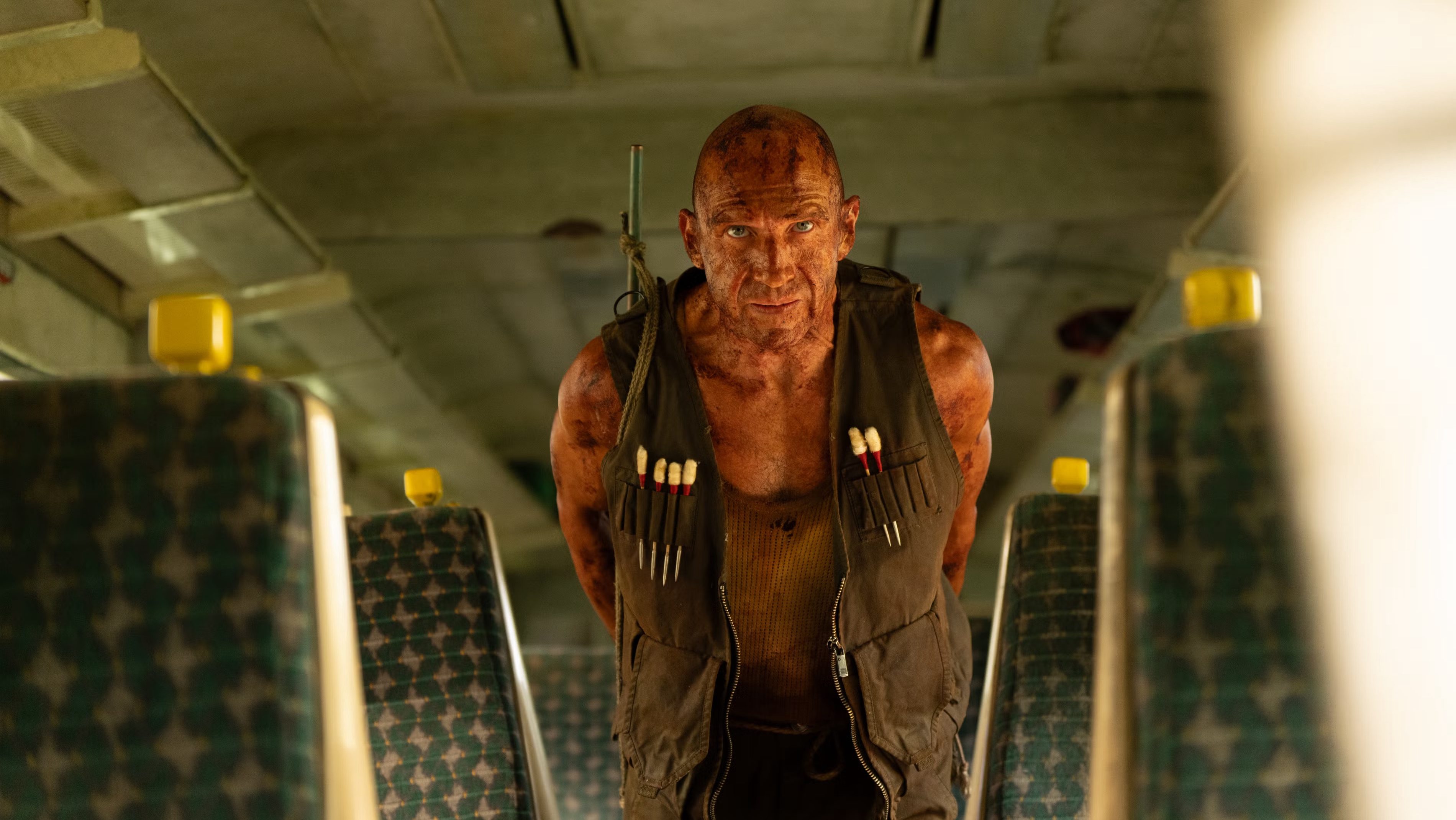 An image from the film 28 Years Later: The Bone Temple. It features a man (Ralph Fiennes) dragging a zombie body through an abandoned train.