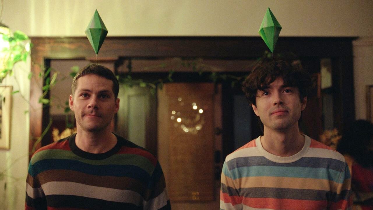 An image from the film Twinless. It features two men (Dylan O'Brien and James Sweeney) wearing colourful striped sweaters, each with a green diamond-shaped SIMS headpiece, standing indoors with a warm ambience.